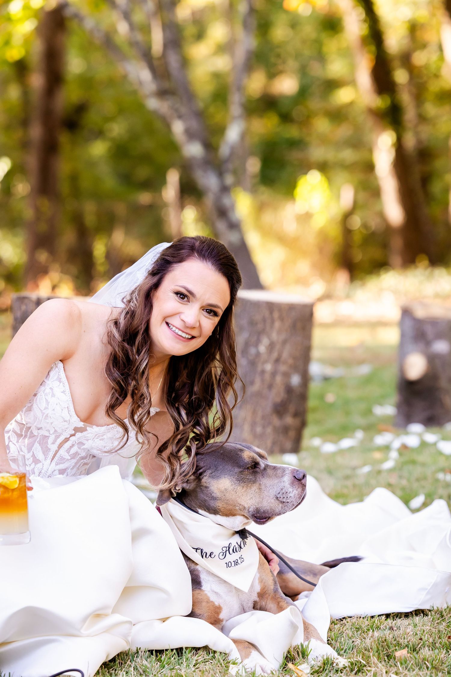 A woman in a white dress relaxes on grass with a pit bull dog during a sunny outdoor photoshoot.