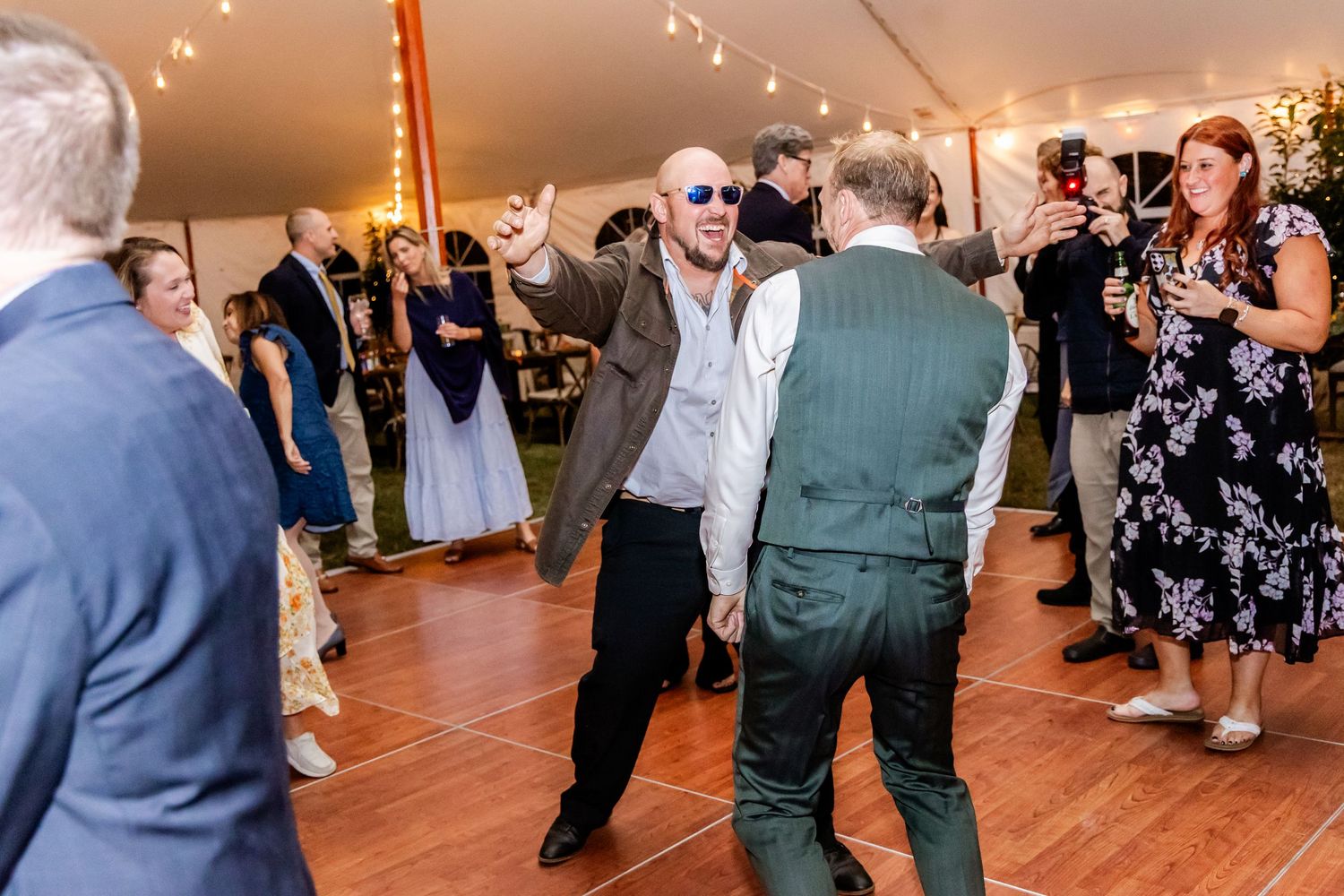 Wedding reception guests dancing on a wooden dance floor under a tent with string lights.