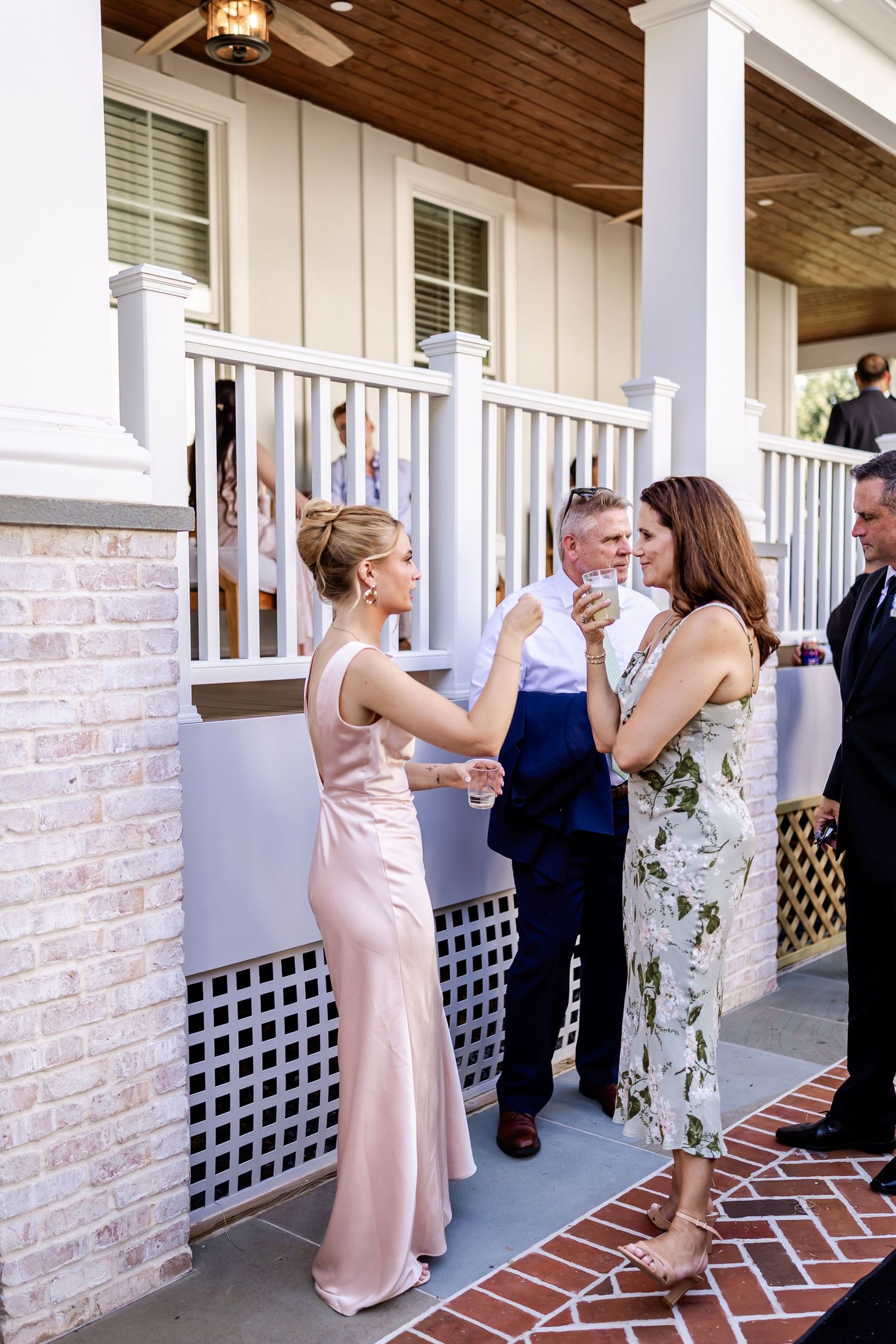Wedding guests socialize and chat on a covered brick porch with white columns and lattice details.