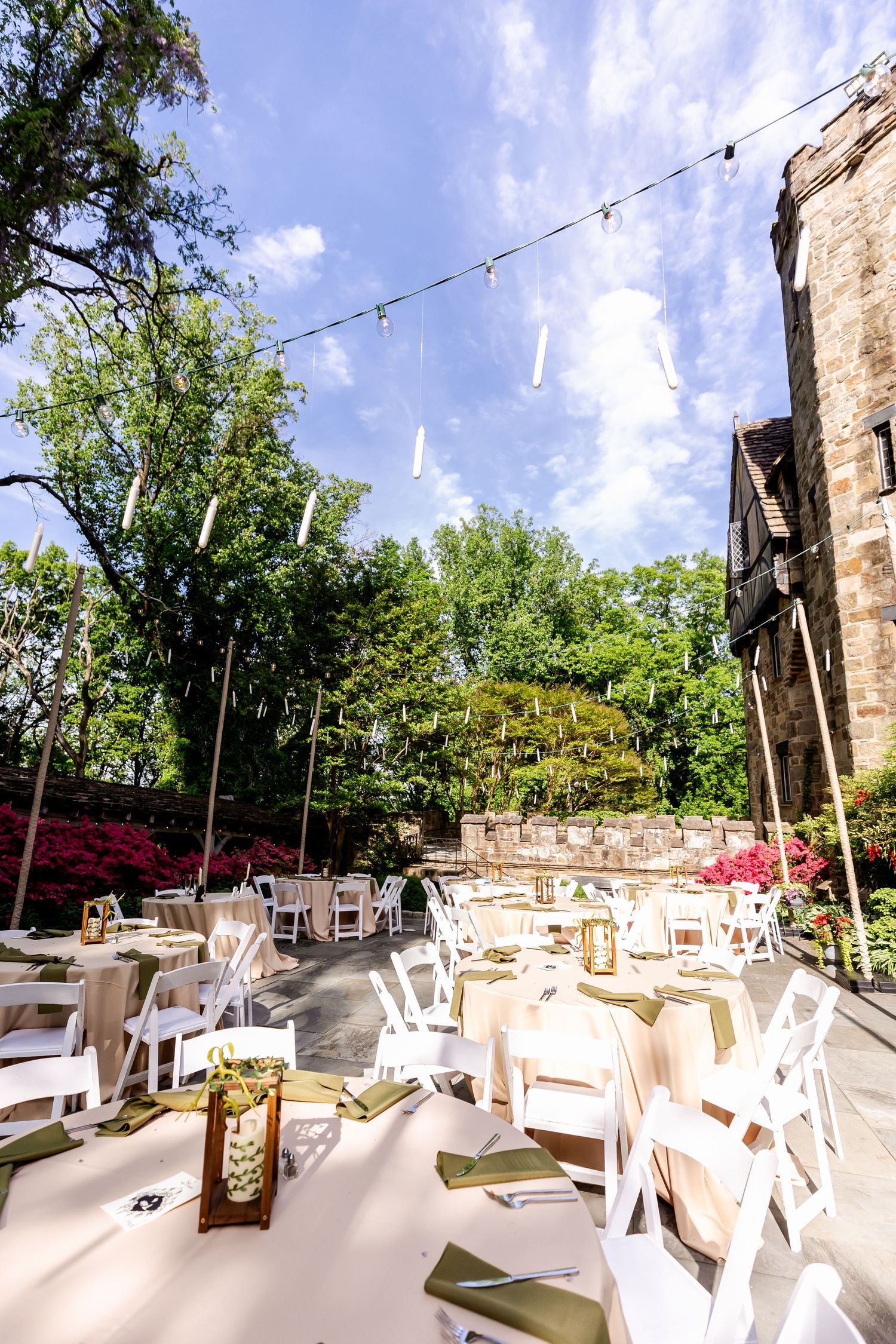 Outdoor wedding reception setup with white chairs and tables under string lights and blue sky.