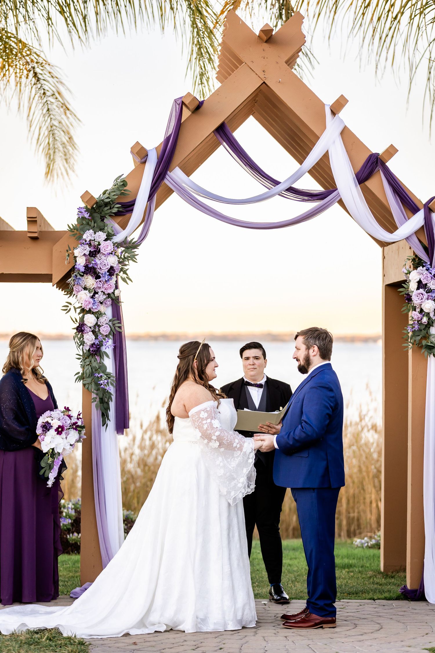 Couple exchanges vows during sunset ceremony under wooden arch with purple and floral decor.