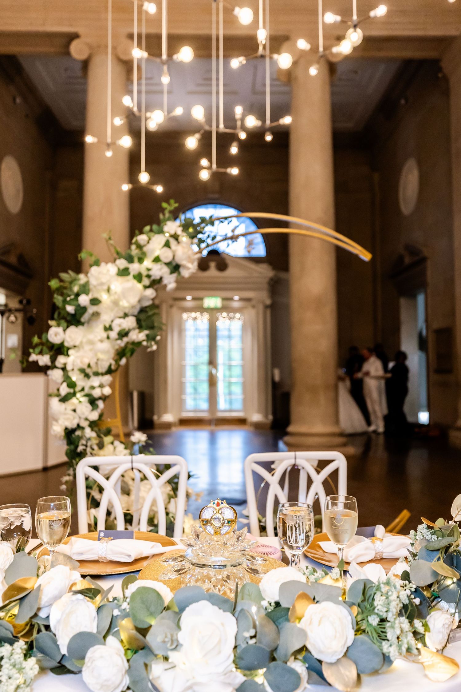 Wedding reception table with gold accents, hanging lights and white floral centerpieces.