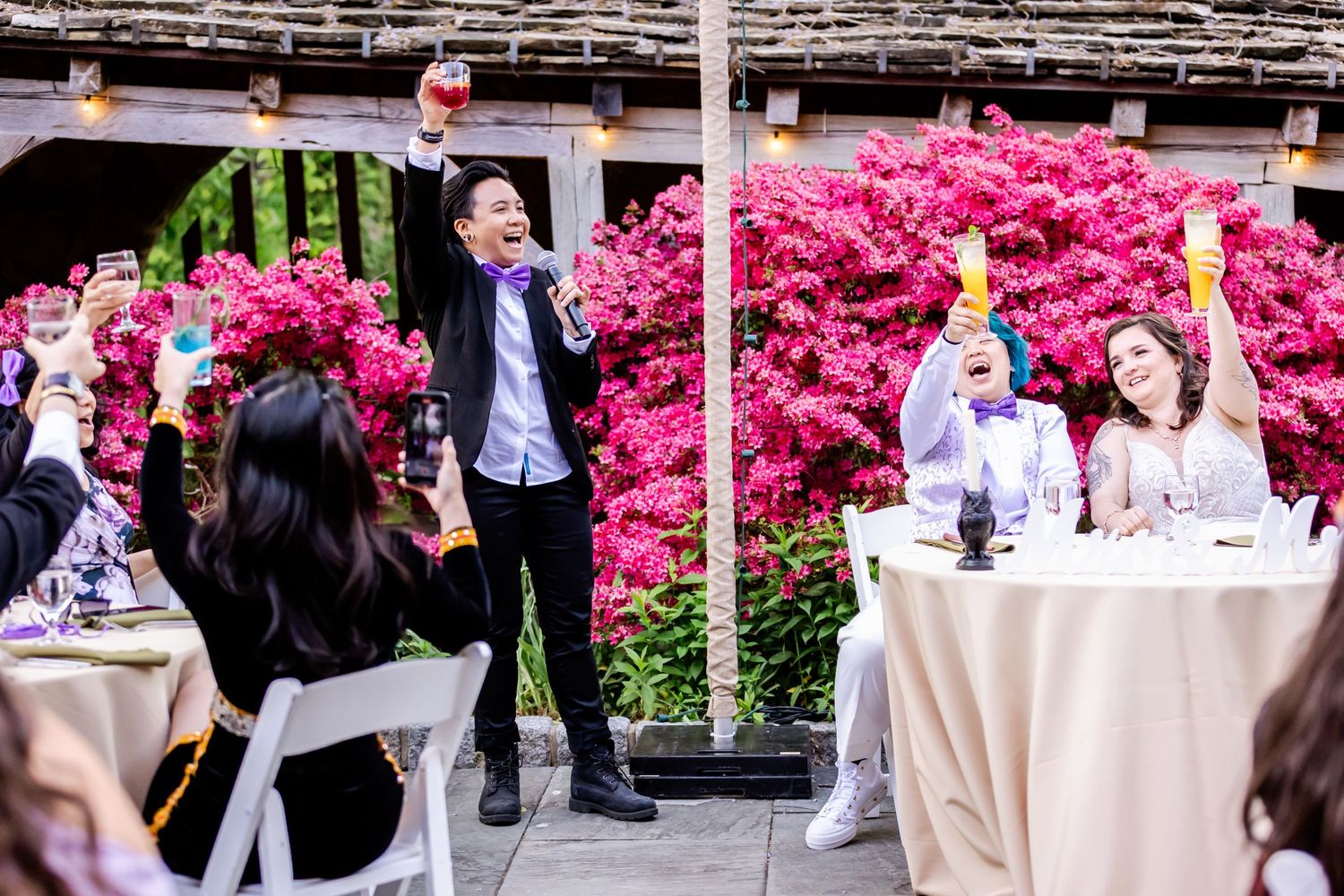Wedding celebration with guests toasting during speeches among pink azalea blooms.