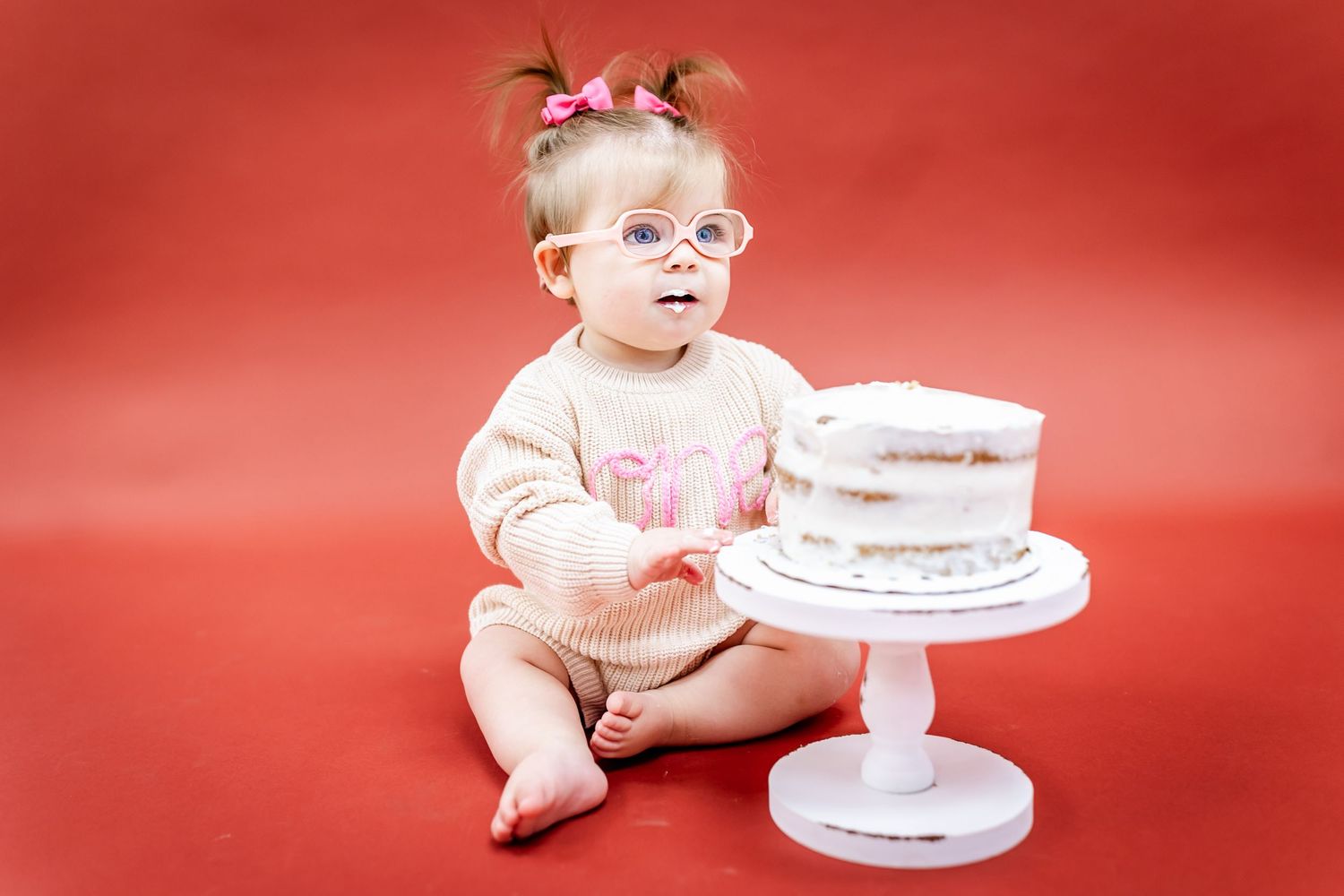 Baby sits beside white cake on pedestal against red background.