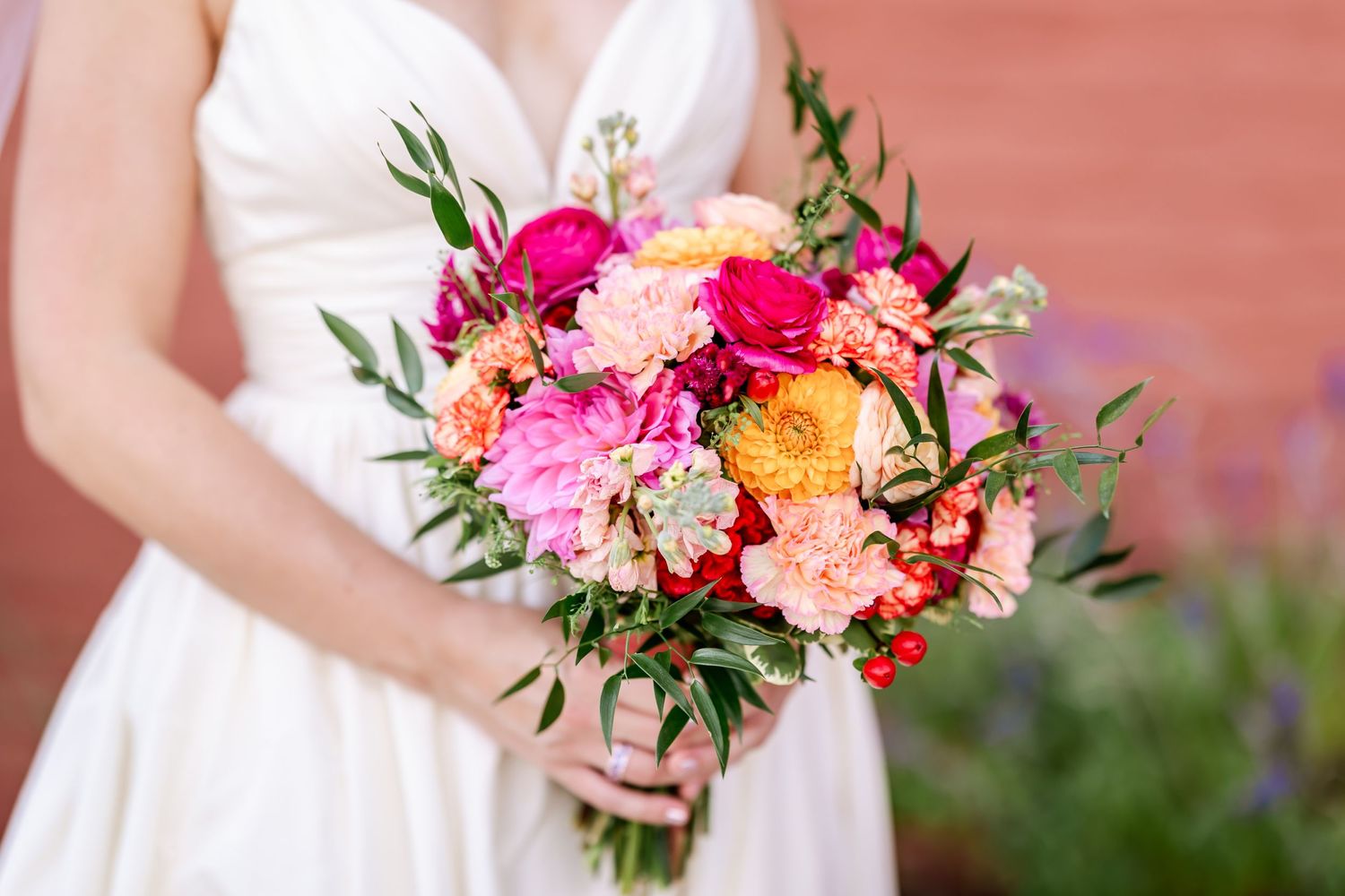 Vibrant bridal bouquet with pink, orange, and coral flowers held against a white wedding dress.
