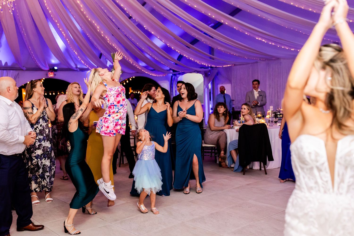 Wedding reception party guests dancing under a purple-lit tent with draped white fabric ceiling at an evening celebration.