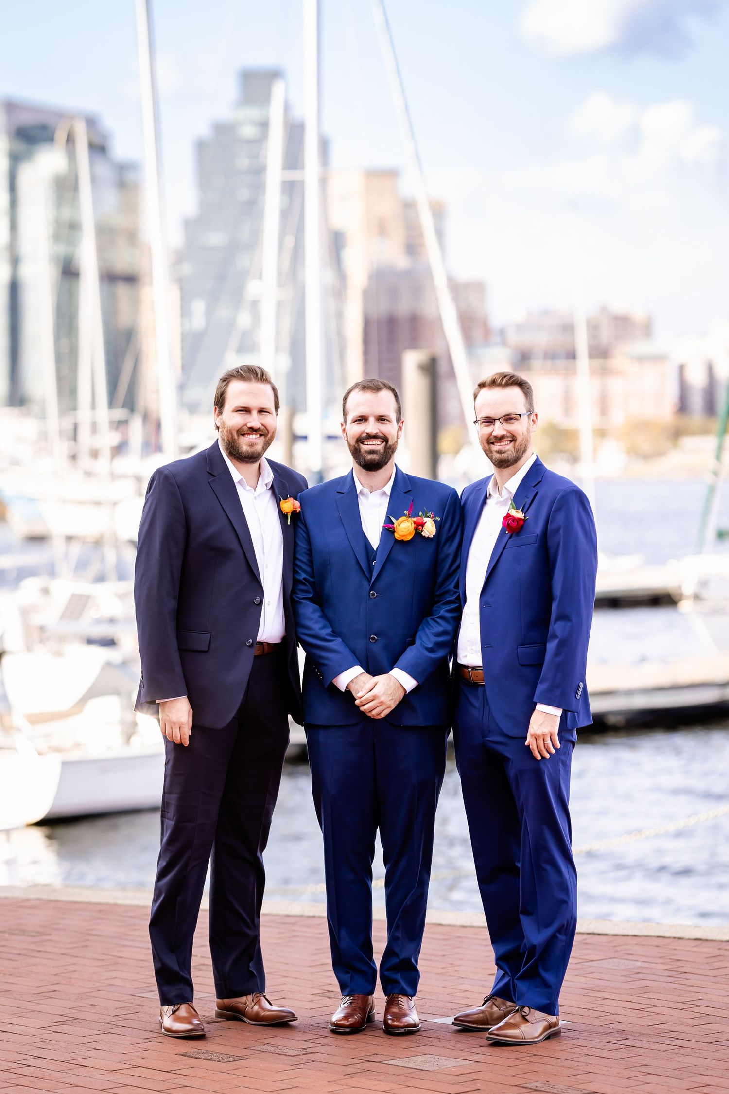 Groomsmen in navy and black suits pose together at a marina with boats and city skyline in background.