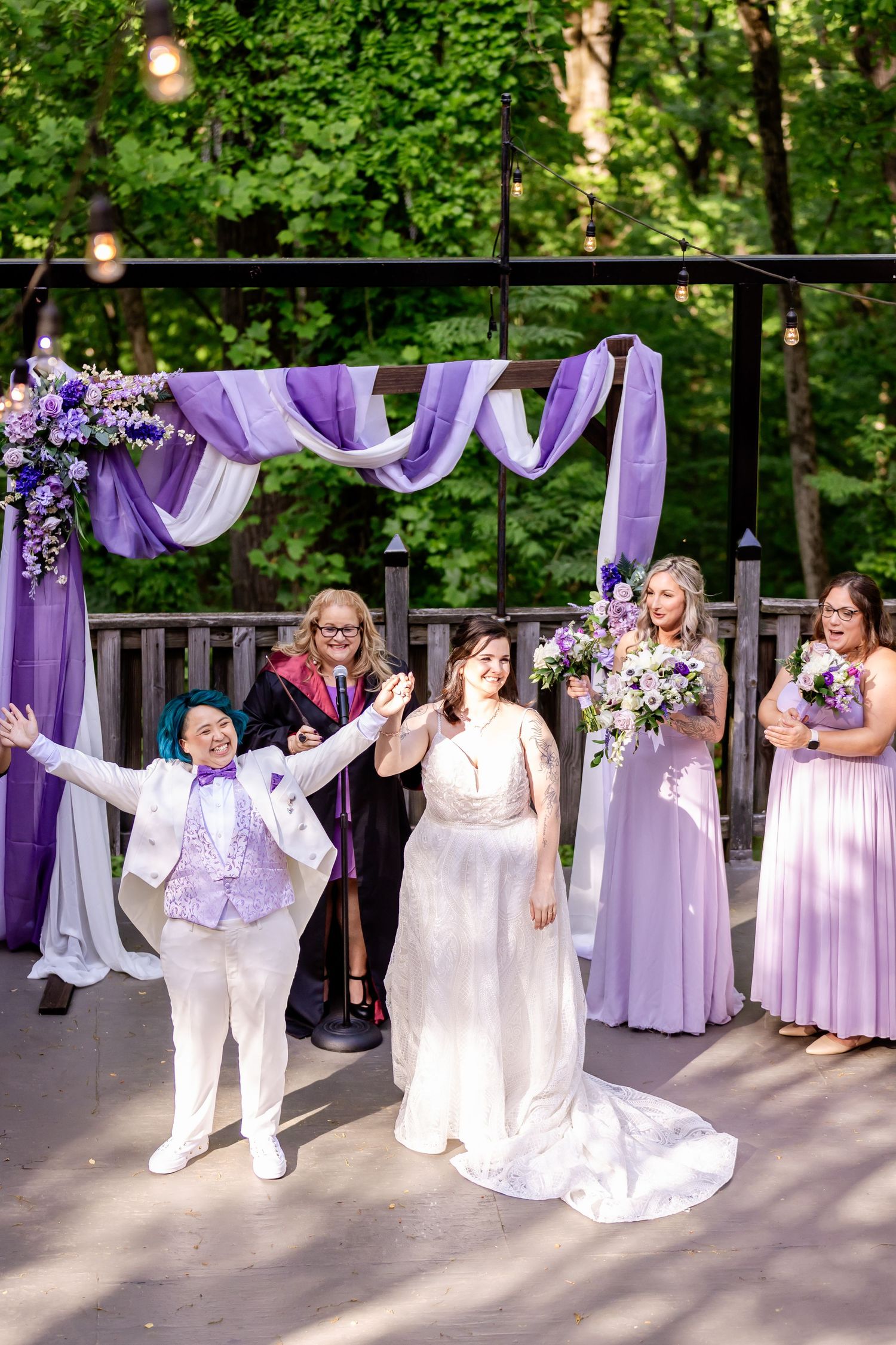 A wedding ceremony under a purple draped arch with wedding party members in lavender dresses outdoors.