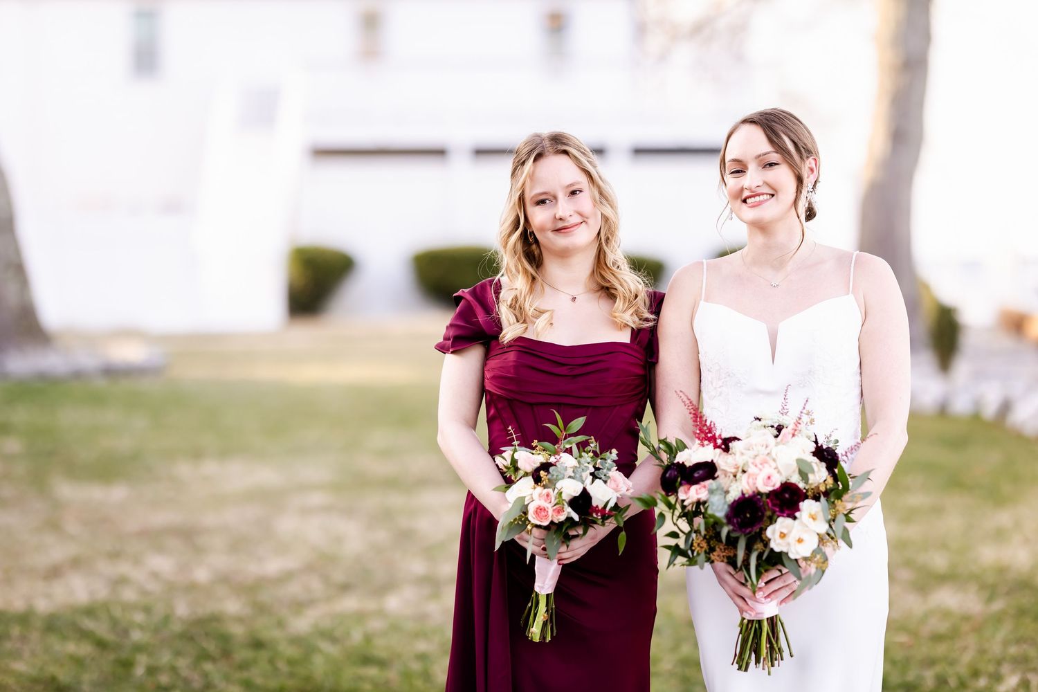 Bridesmaids in burgundy dresses hold bouquets while standing together outdoors.