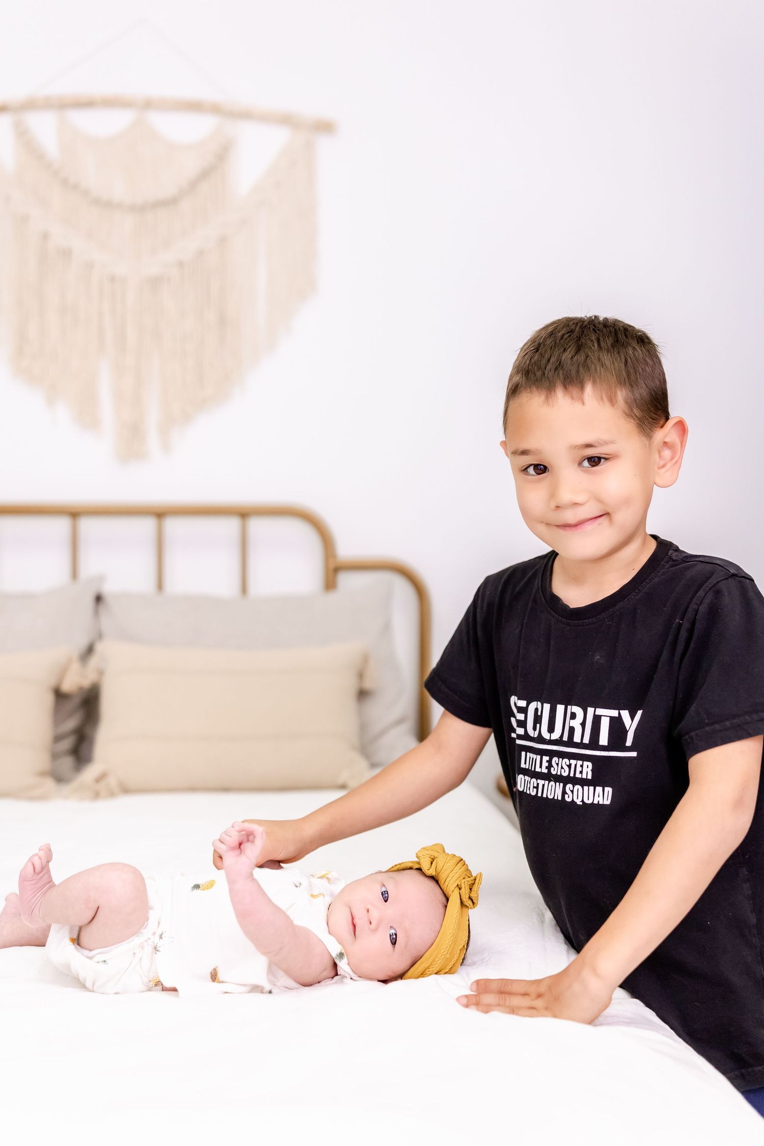 Two children on a bed with one wearing a black security shirt and decorative wall art in the background.