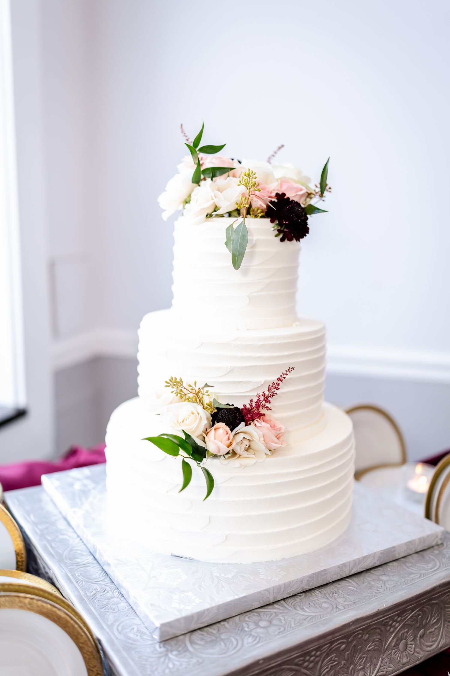 Elegant white tiered wedding cake decorated with pink flowers and greenery on a marble stand.