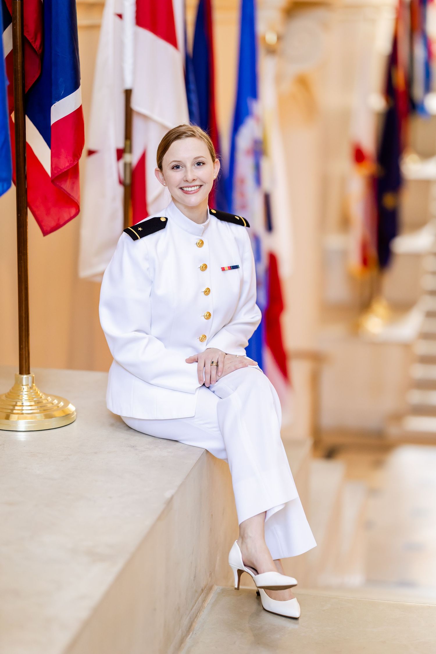 Naval officer in dress whites sits elegantly on ledge with international flags displayed in background.