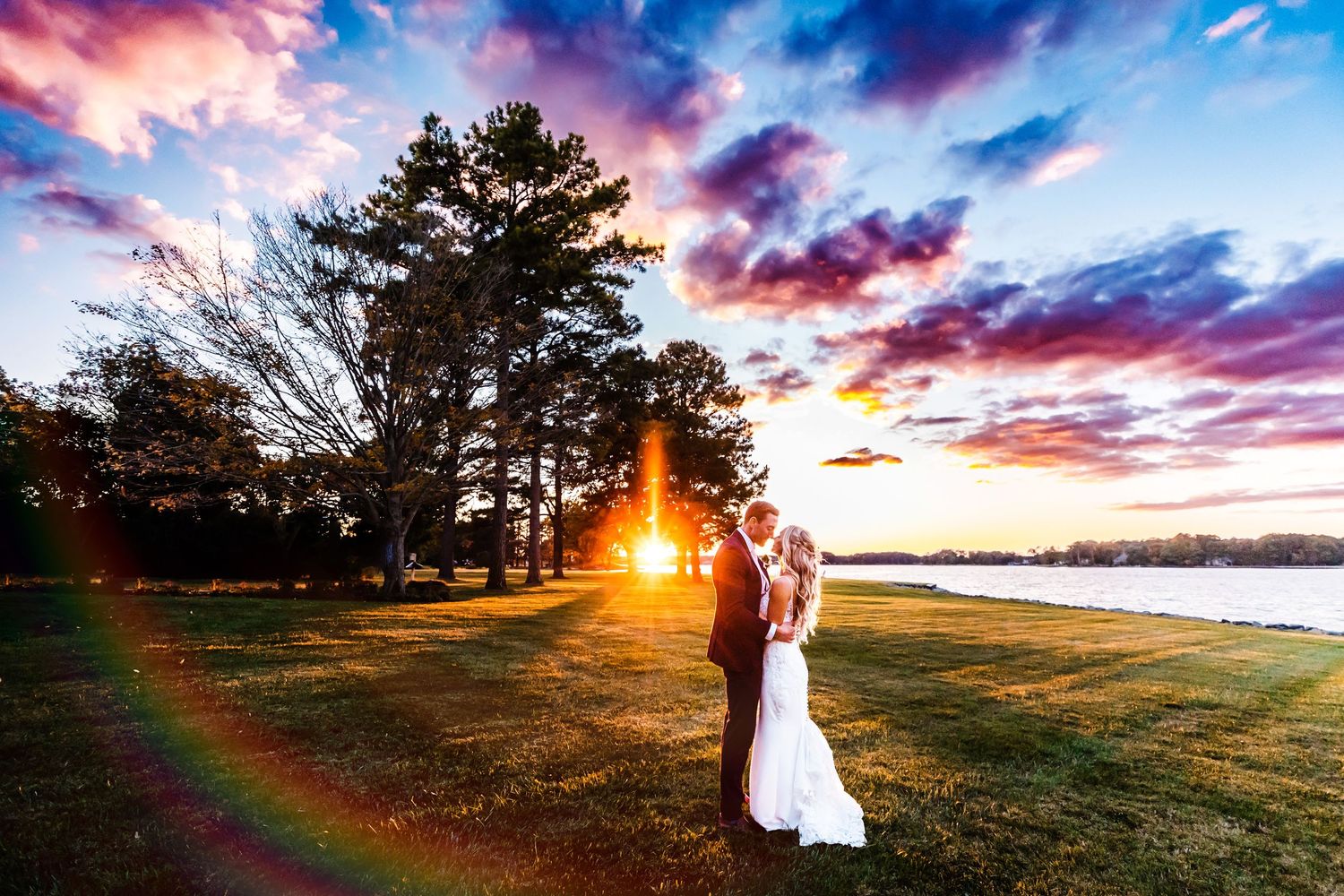 Stunning sunset silhouettes couple on lakeside lawn with dramatic pink and purple clouds in background.