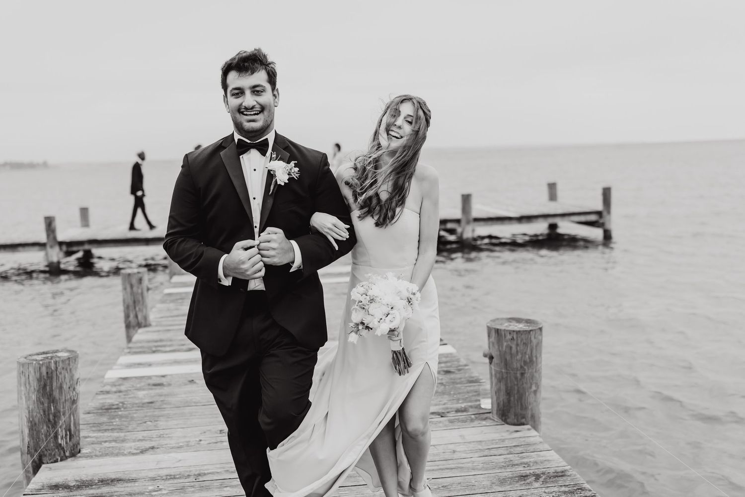 Wedding couple walking and laughing together on a beach with wooden posts in the background.