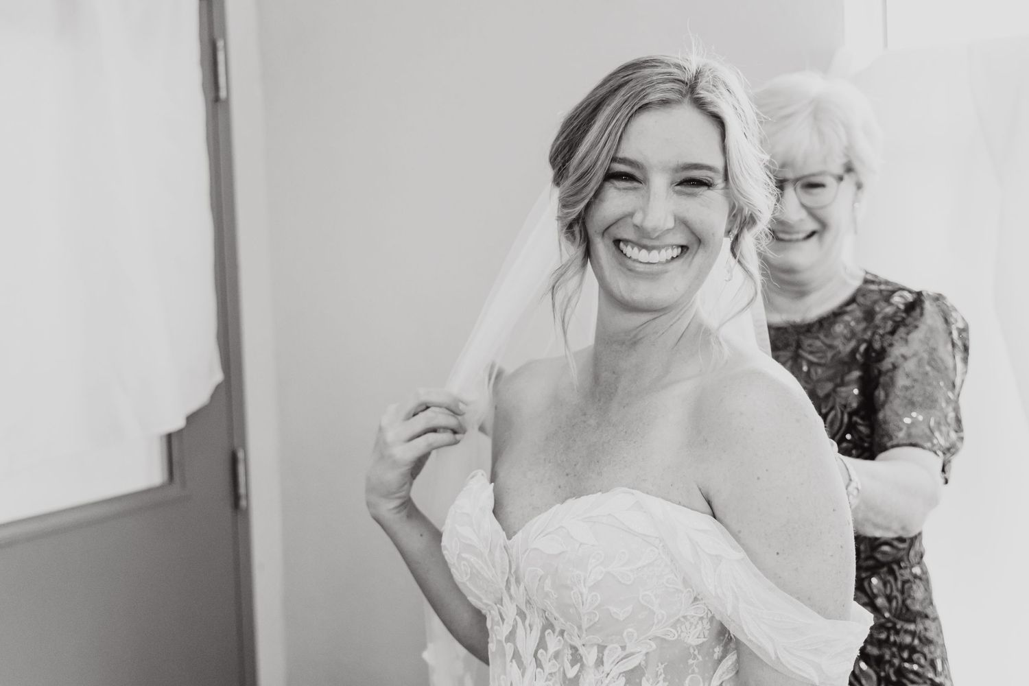 Black and white photo of a joyful moment showing a strapless gown fitting with two people in a bridal preparation room.