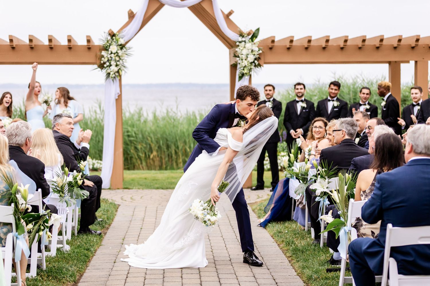 Wedding party poses outdoors with white floral decorations and wooden pergola backdrop.