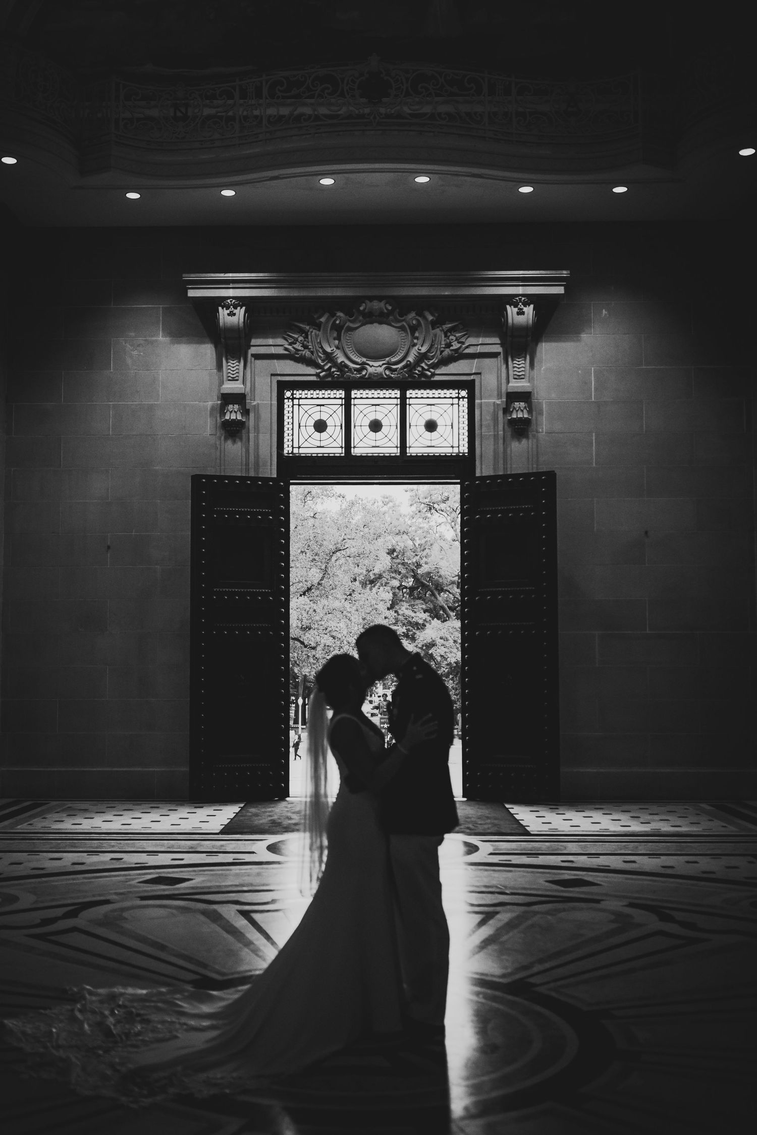 Silhouetted couple in formal attire embraces in dramatic backlit doorway of grand building entrance.