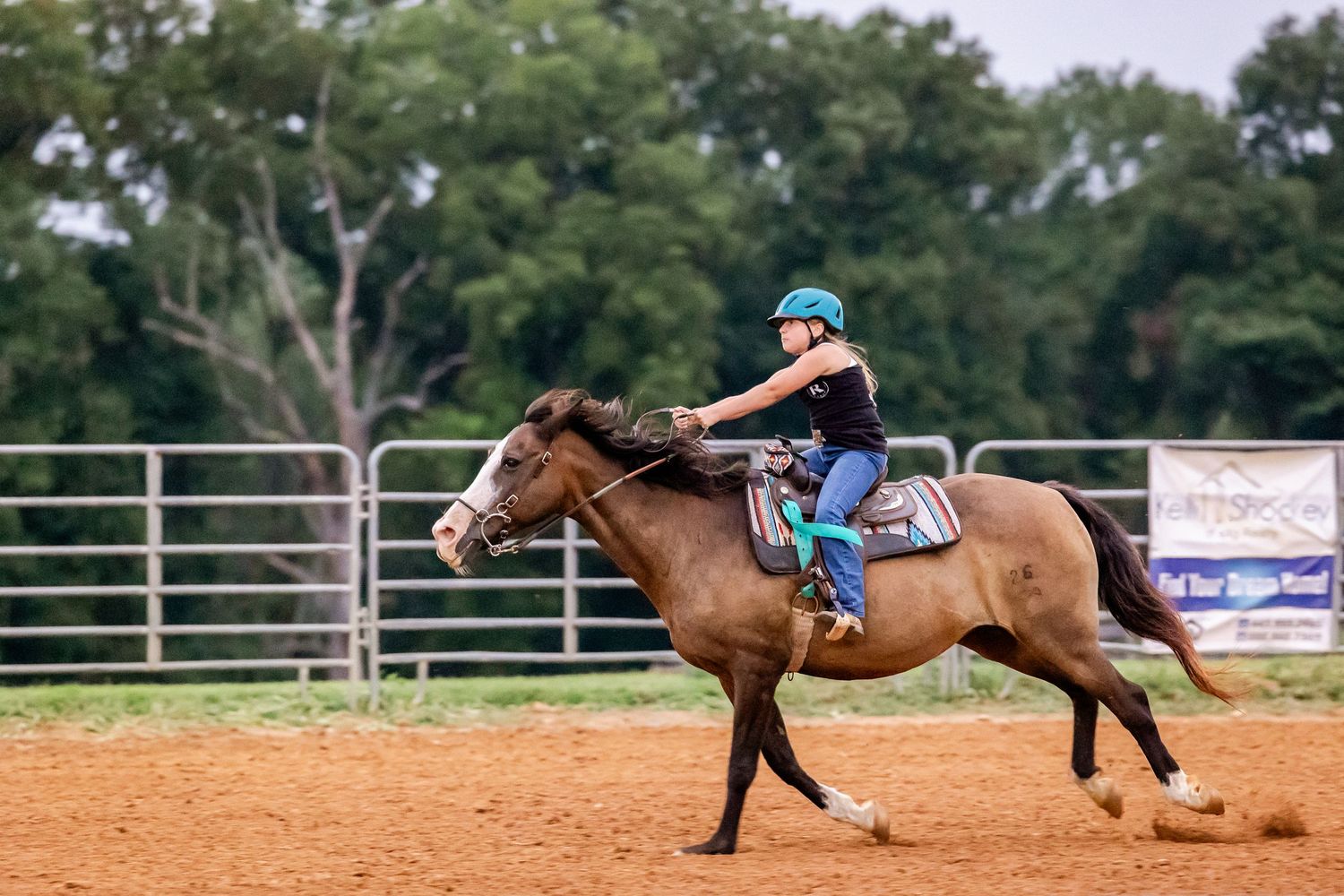 Young rider in blue practices horseback riding in a sandy arena with metal fencing.