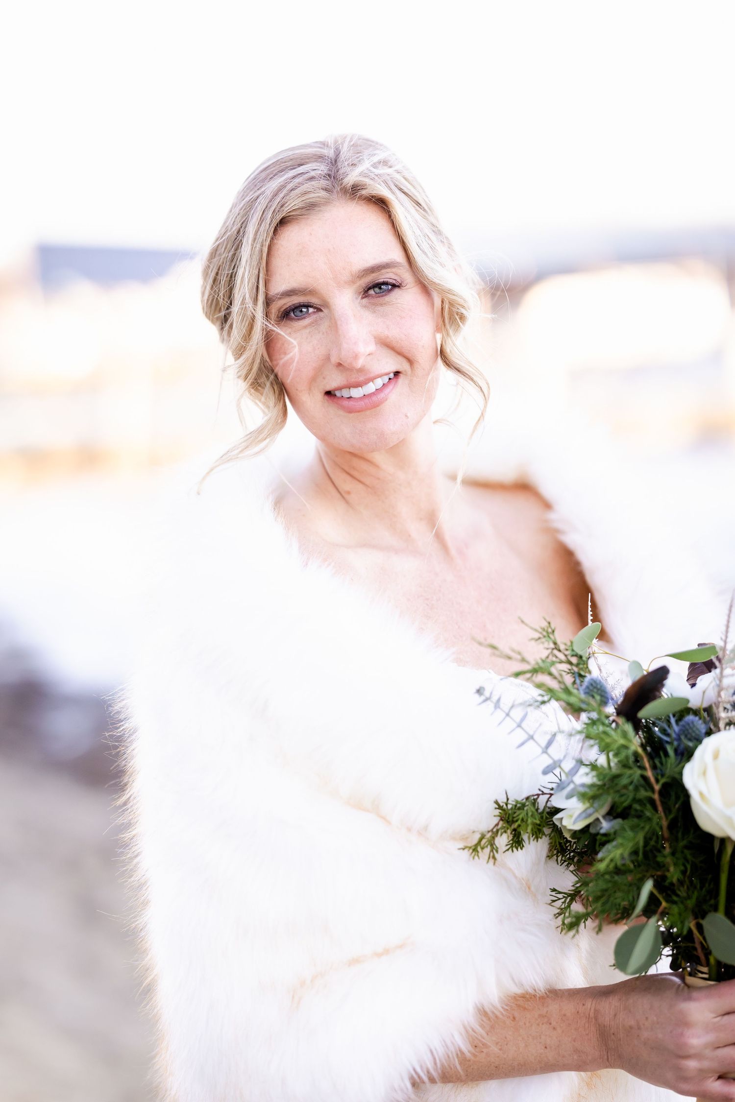 Winter bride in white fur wrap holds evergreen bouquet against bright snowy backdrop.