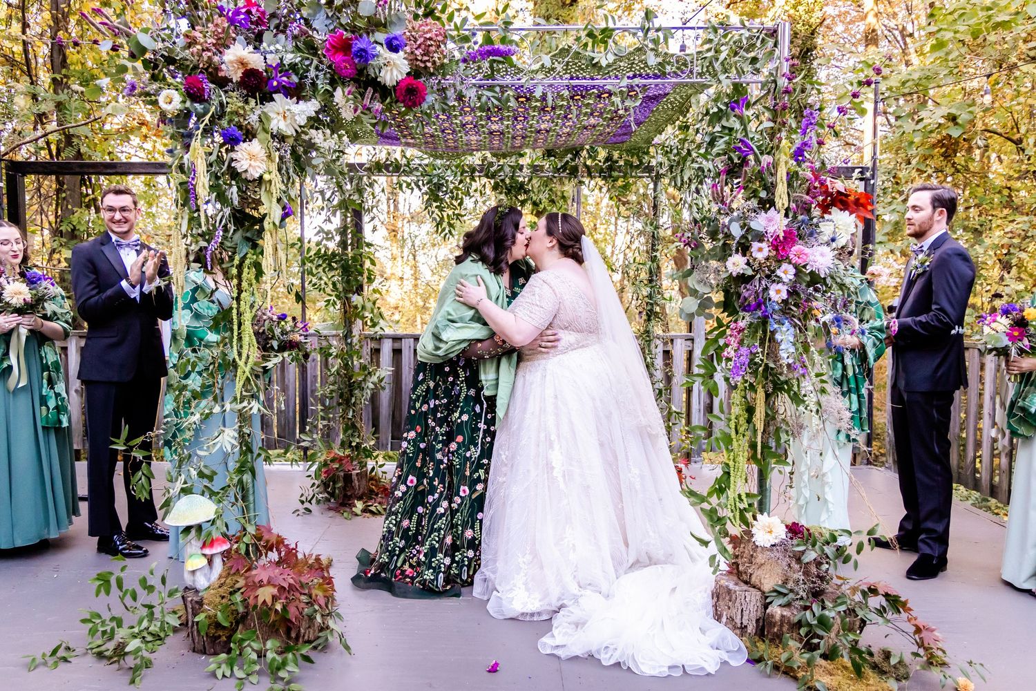 Wedding ceremony under floral arch with purple and pink blooms in garden setting.