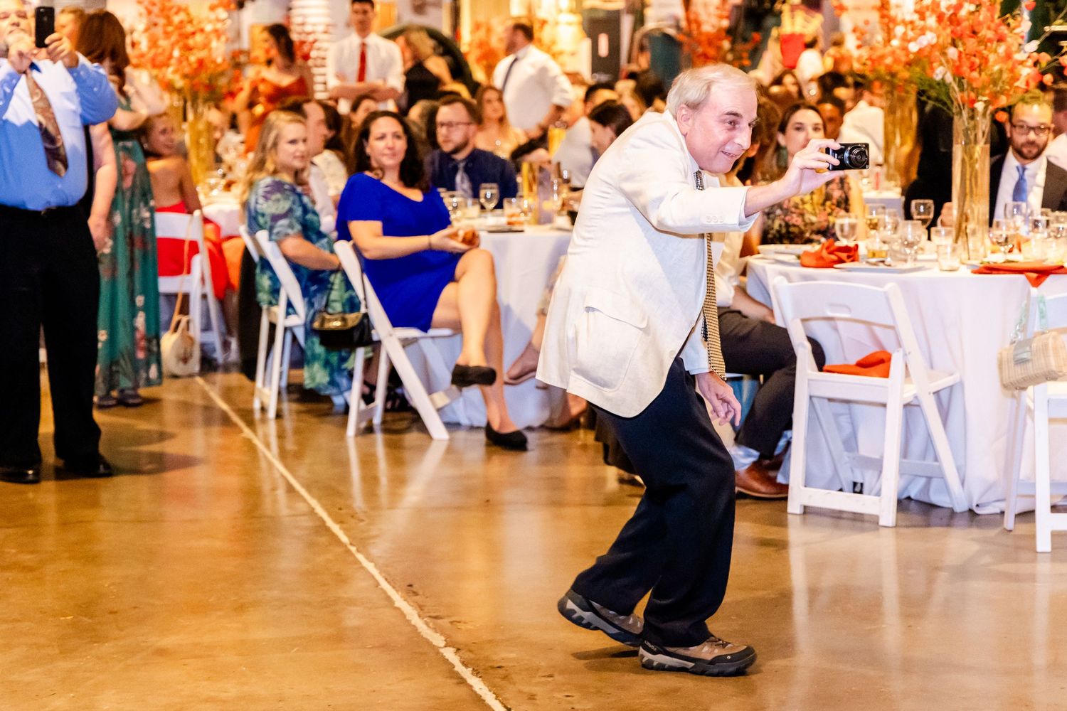 Wedding guest performs a limbo dance move at a reception while other guests watch from decorated tables in the background.