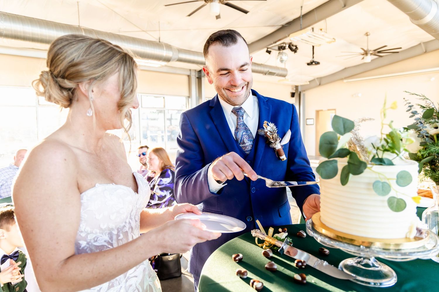 A couple in wedding attire cut their green and white tiered wedding cake at an indoor reception.