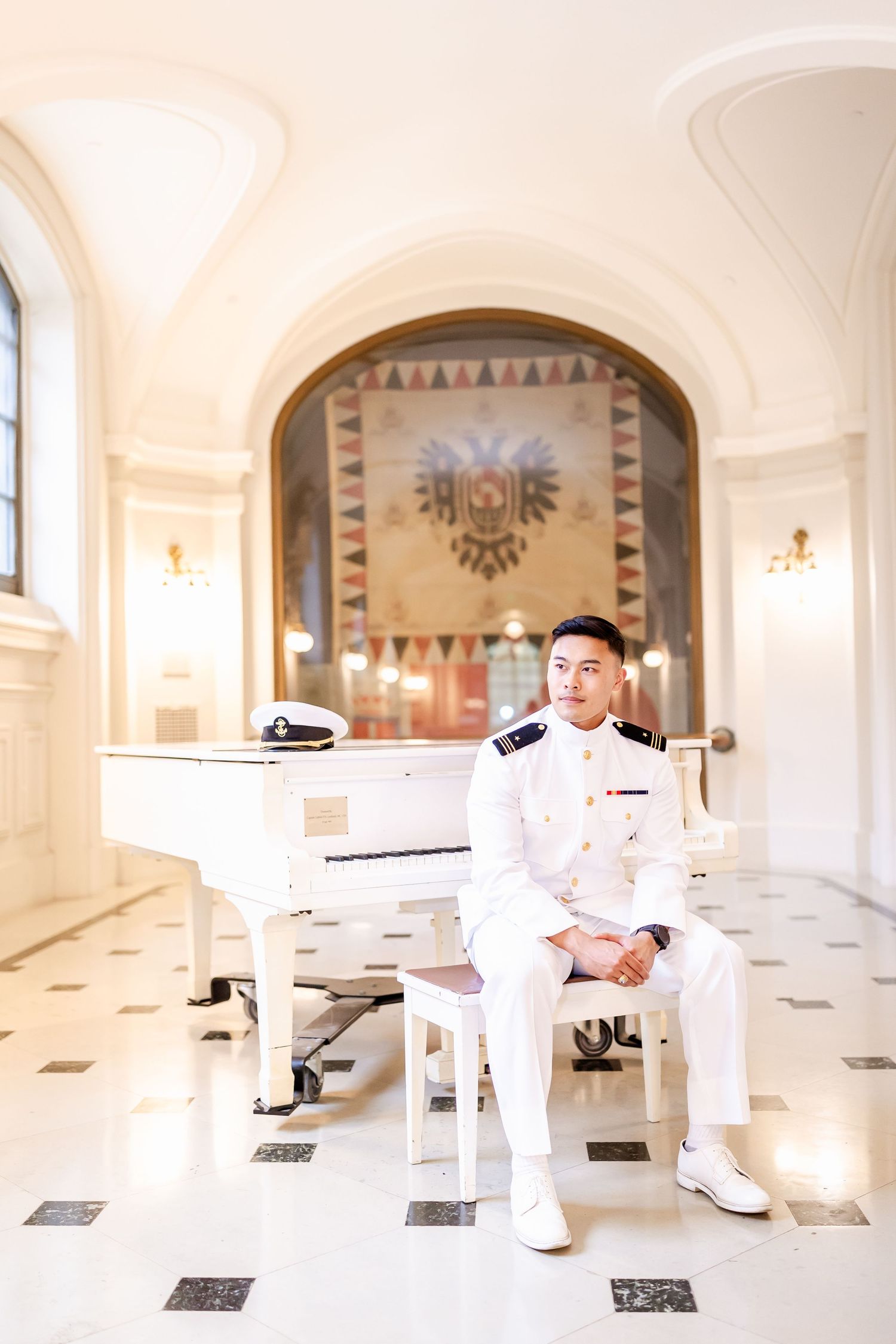 A person in a white naval uniform sits at a grand white piano in an elegant room with arched ceilings and marble floors.