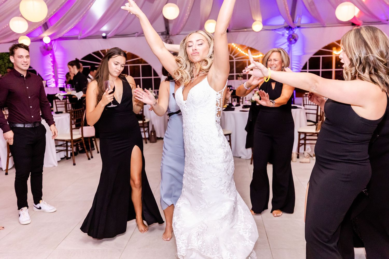 Bride dancing with guests at wedding reception under purple lighting and white tent.