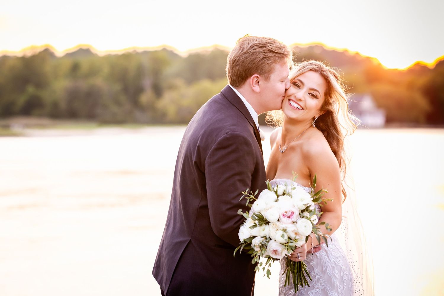A romantic wedding couple embraces during sunset by a lake holding a pink and white floral bouquet.