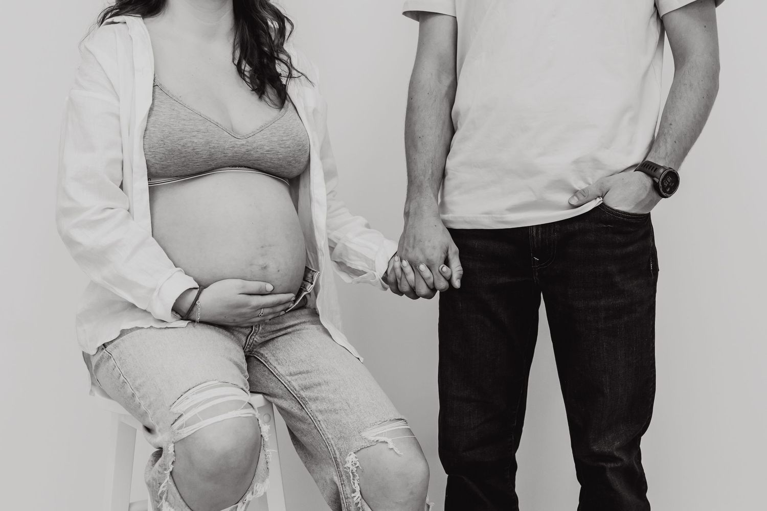 Black and white photo showing couple holding hands with expectant mother wearing ripped jeans and white top.