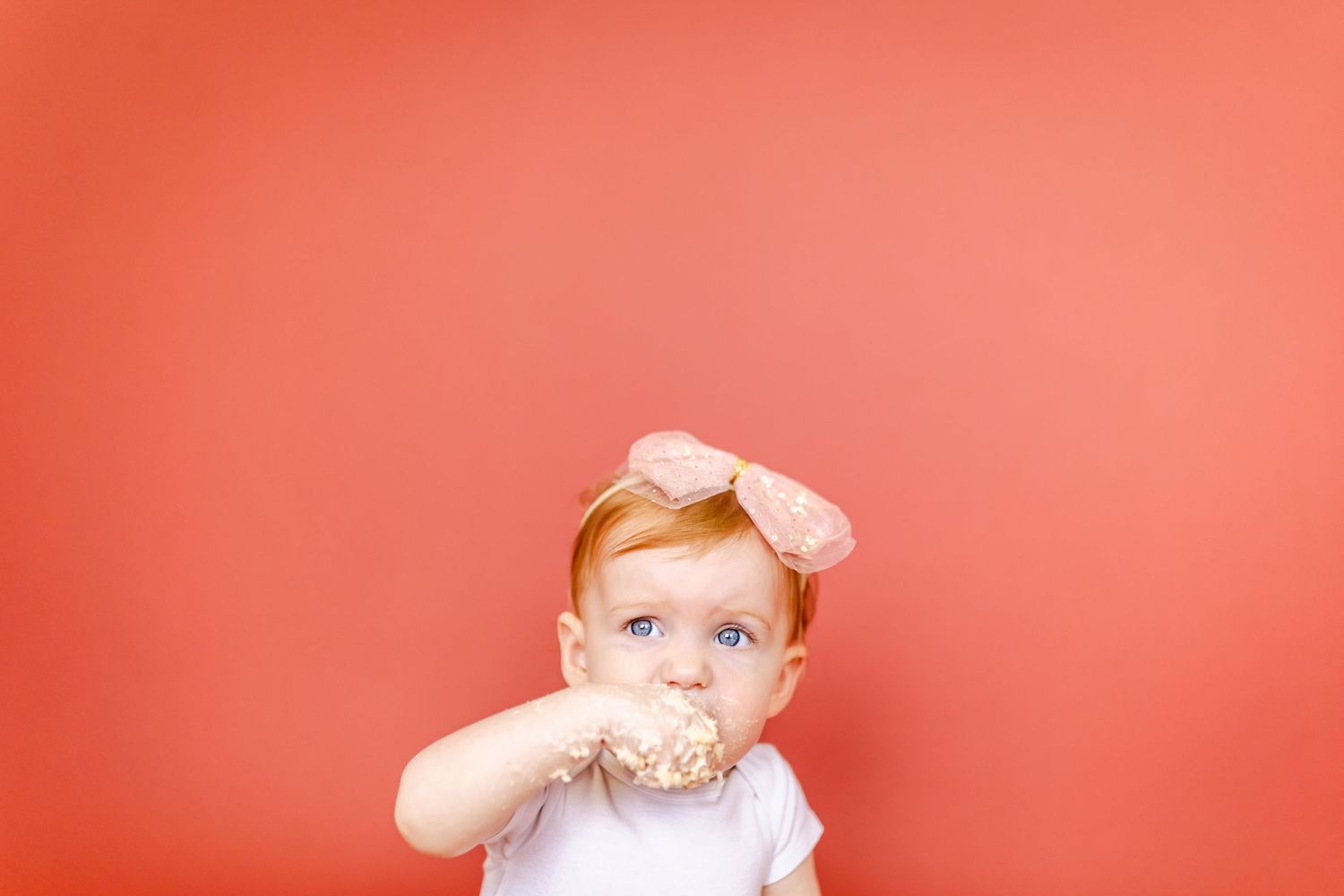 Baby in pink dress plays on white chair with coral colored background.
