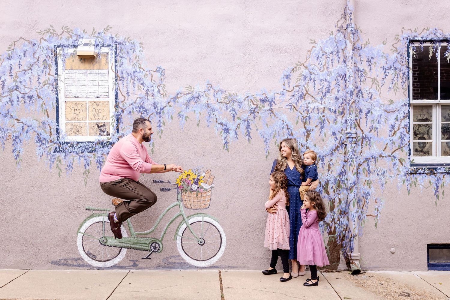 Family walking and playing along a purple floral wall with a vintage bicycle.