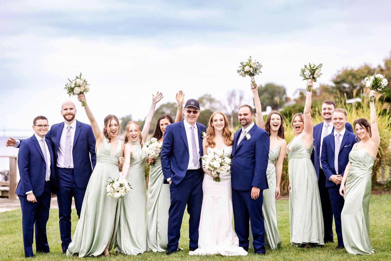 Wedding party in navy suits and sage green bridesmaid dresses pose together outdoors on a sunny day.