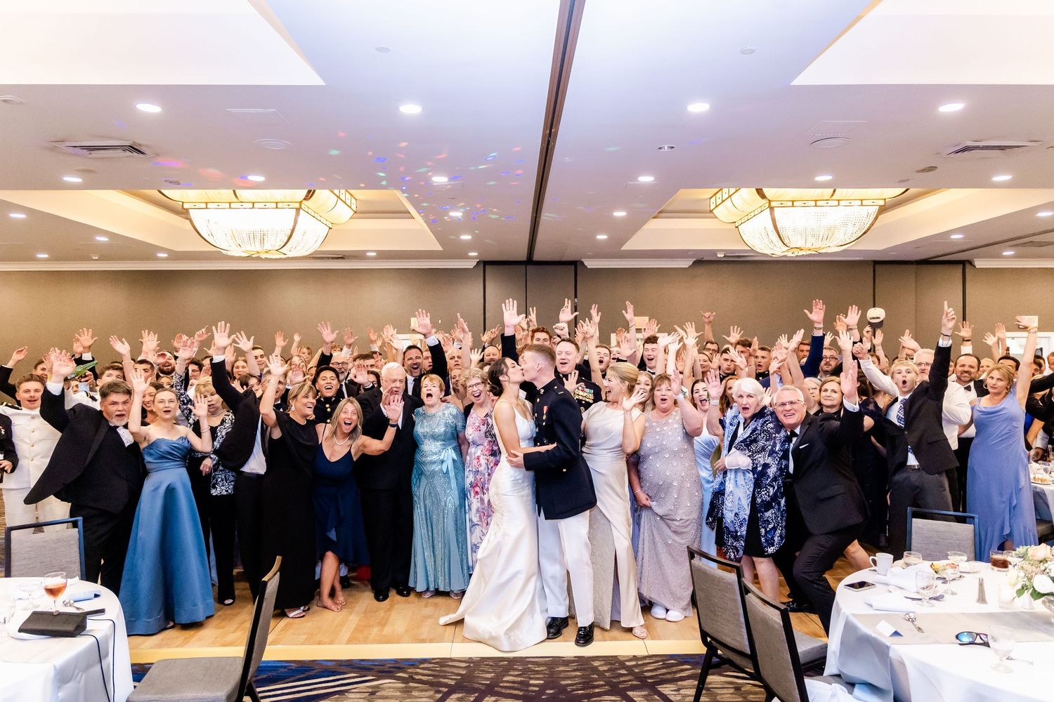 Large group of wedding guests celebrating in an elegant ballroom with crystal chandeliers.