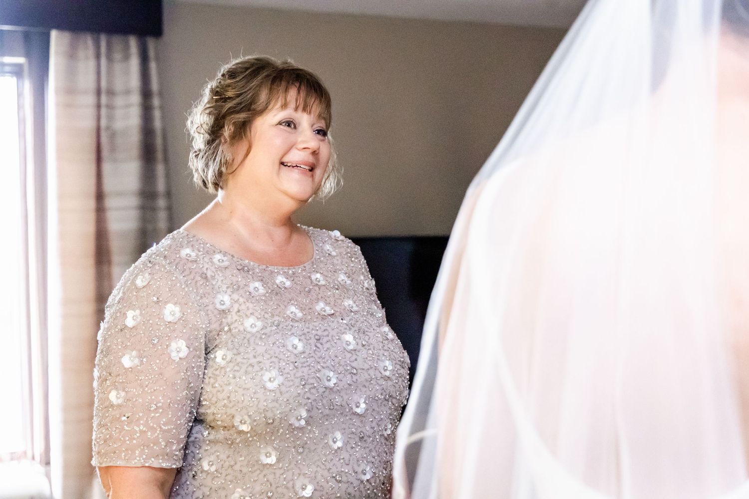A bride in a grey beaded dress smiles while holding her veil.