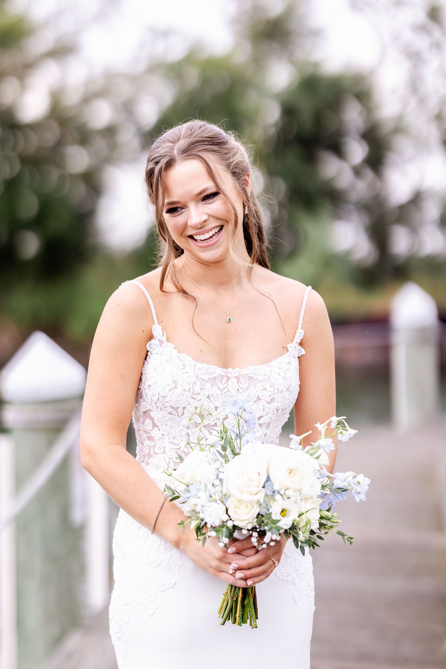 Joyful bride in lace dress holds white floral bouquet during outdoor wedding photos.