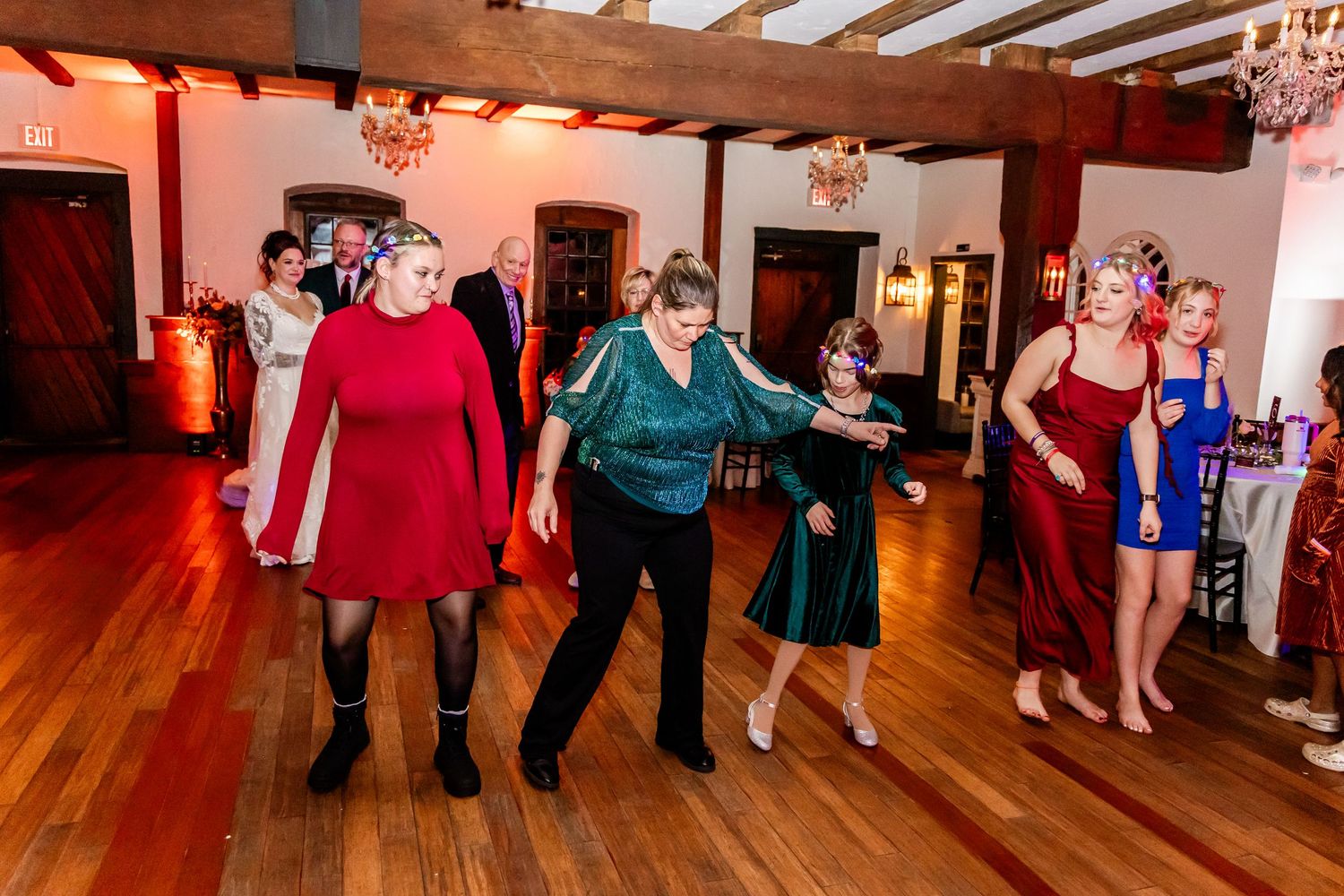 People dancing together on a wooden dance floor at an indoor event with red ambient lighting and exposed ceiling beams.