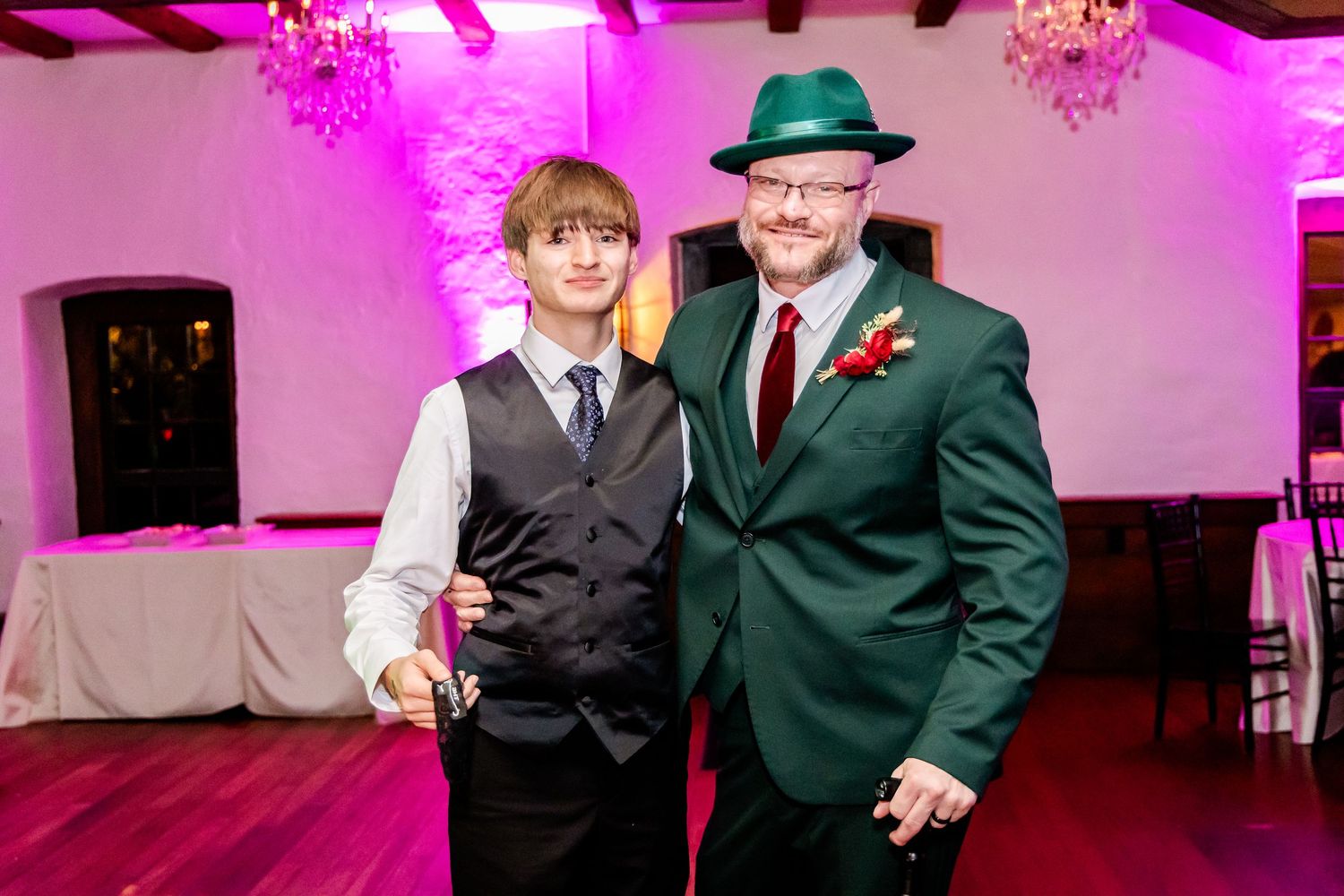 Two guests pose together at a wedding reception under pink lighting with one wearing a green suit and hat.