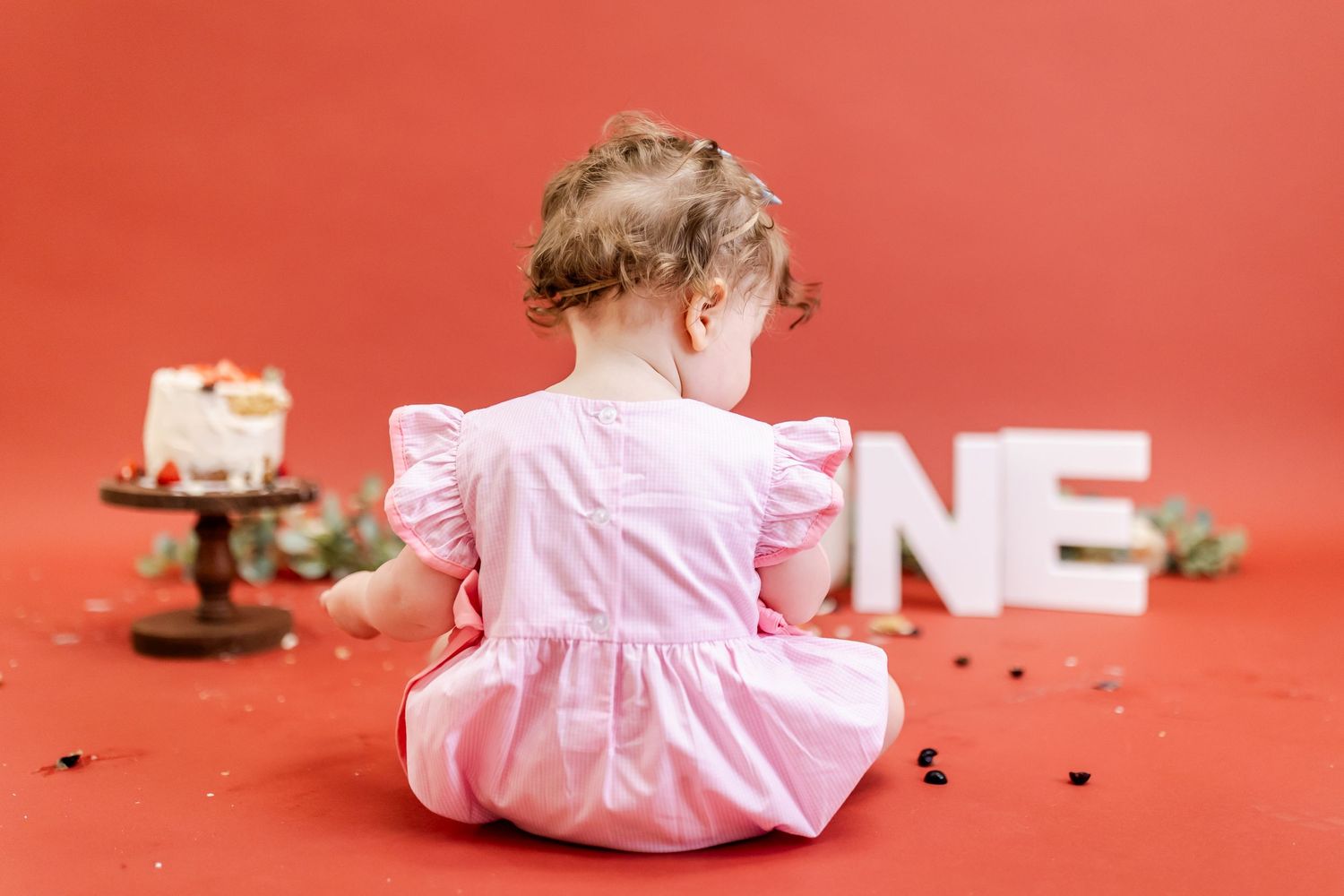 A baby in a pink dress sits on a red floor while playing with scattered toys and objects.