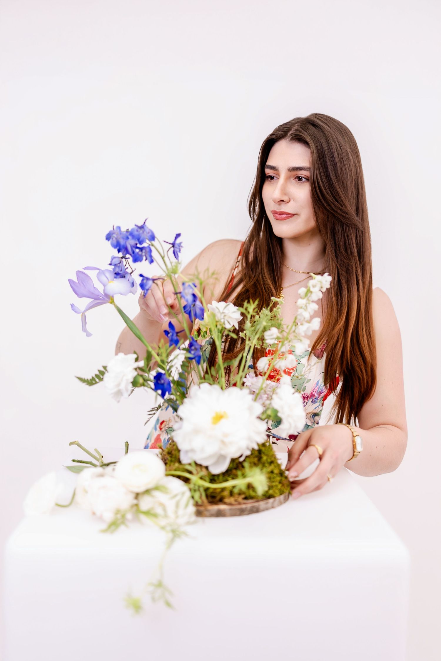 Young woman with long brown hair arranging blue and white flowers in a rustic basket on white table.