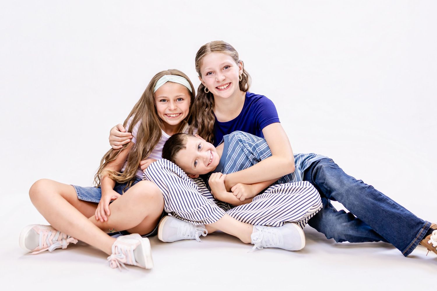 Three happy kids sitting together on white background, two girls hugging a boy, wearing casual jeans and striped outfits.