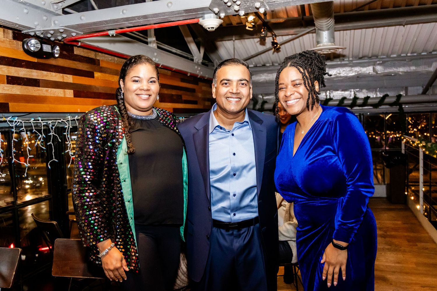Three people in formal attire posing together at an elegant event venue with industrial ceiling.