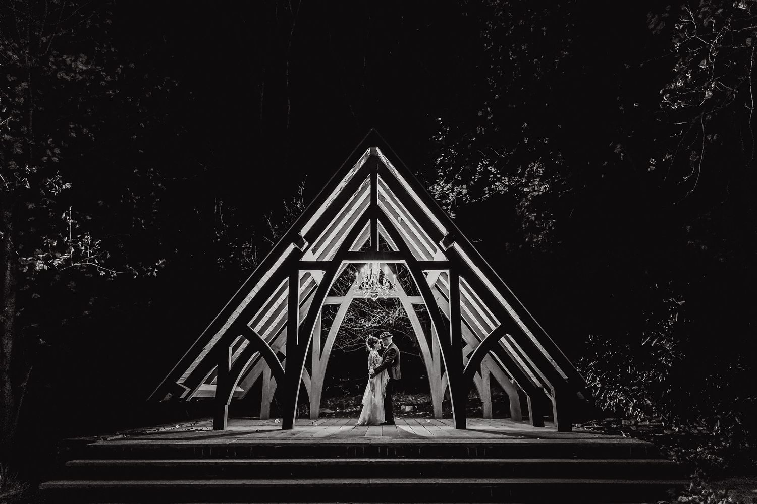 A dramatic black and white wedding photograph of a couple silhouetted under a triangular wooden chapel structure.
