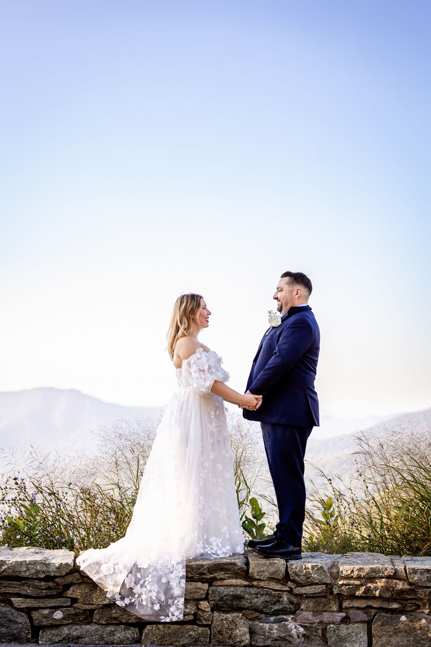 A couple in wedding attire shares romantic moments along a stone wall with mountain views in the background.