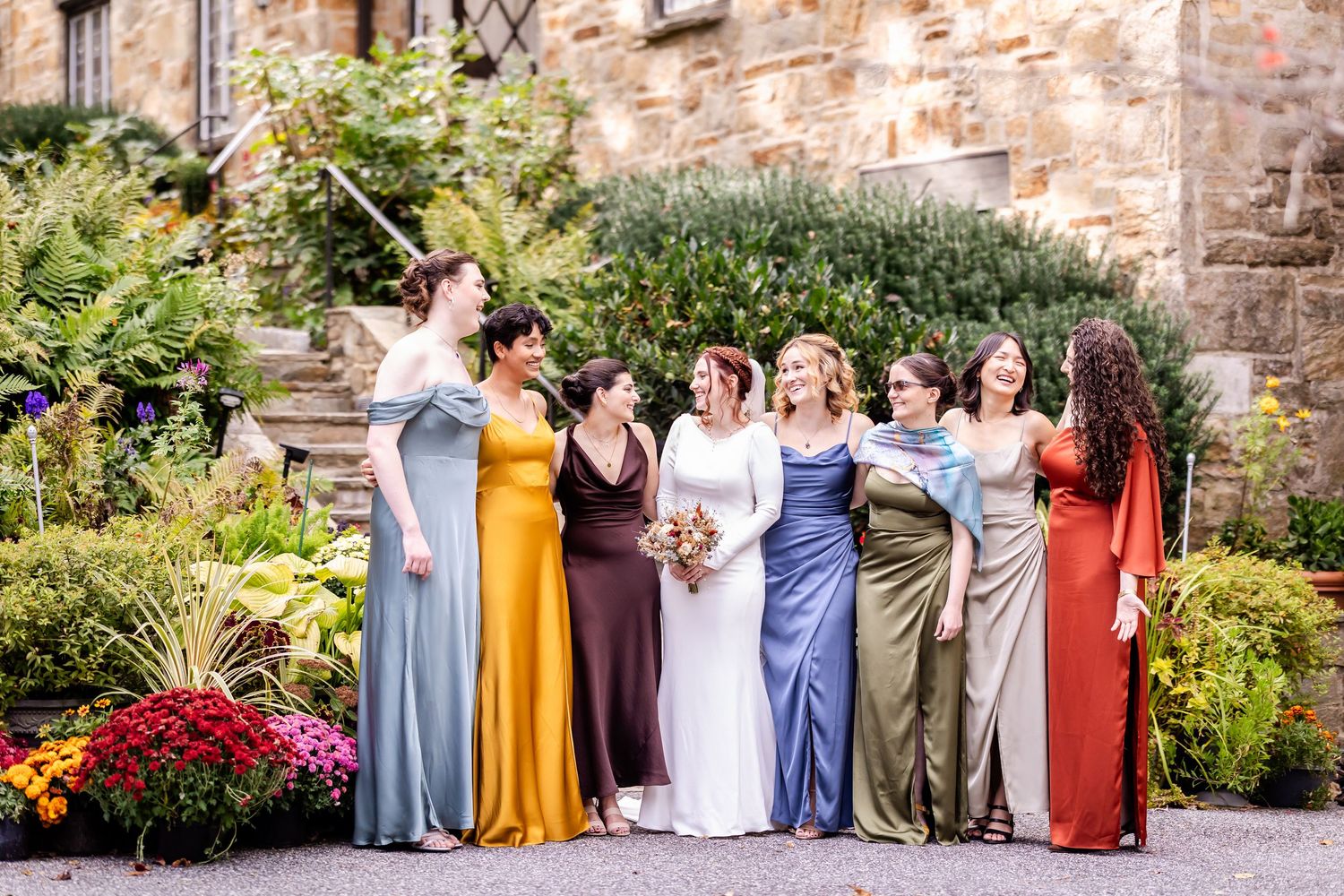A group poses together in formal attire against a rustic stone wall backdrop with colorful garden flowers.