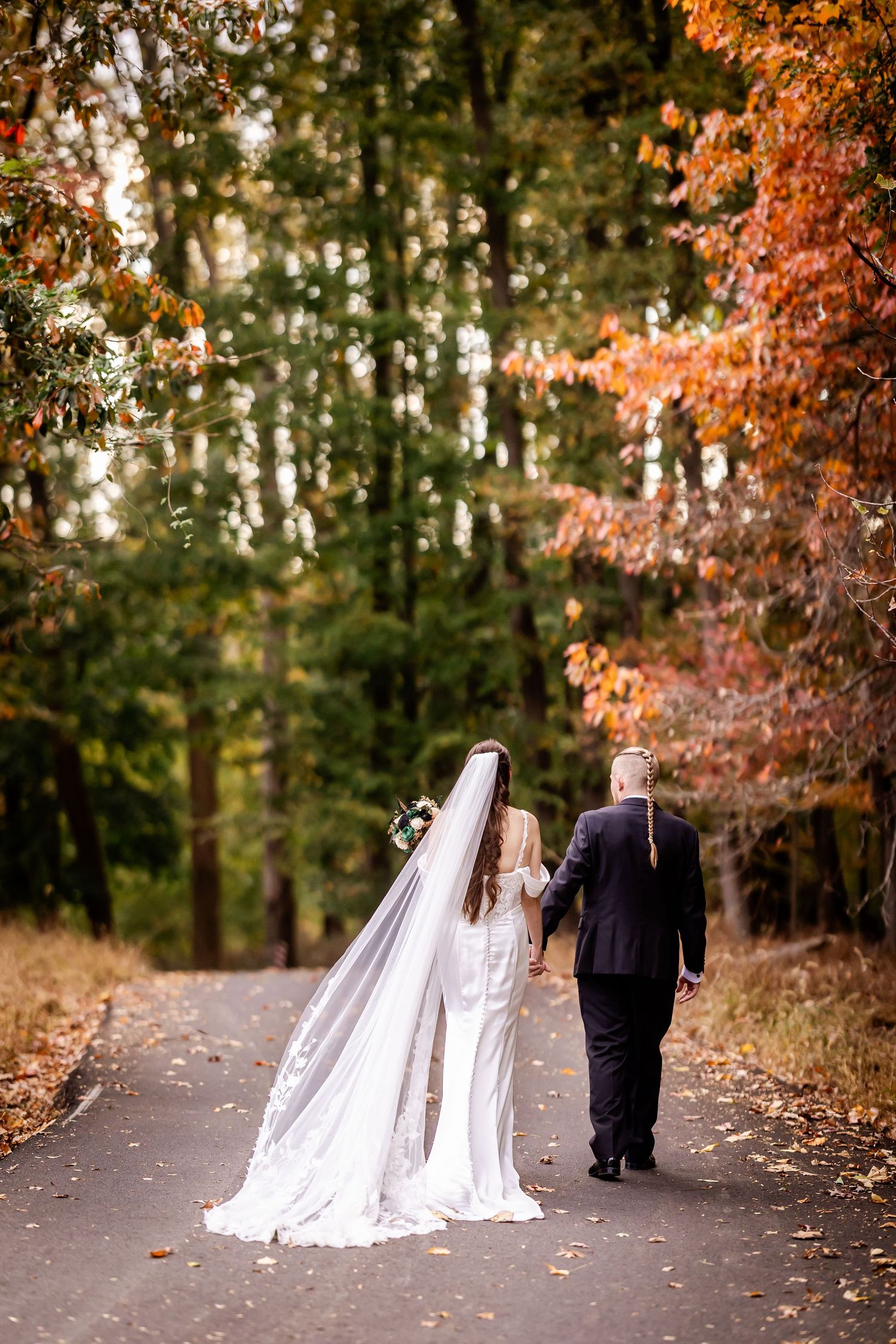 A couple in wedding attire walk down a tree-lined path surrounded by autumn foliage and golden sunlight.
