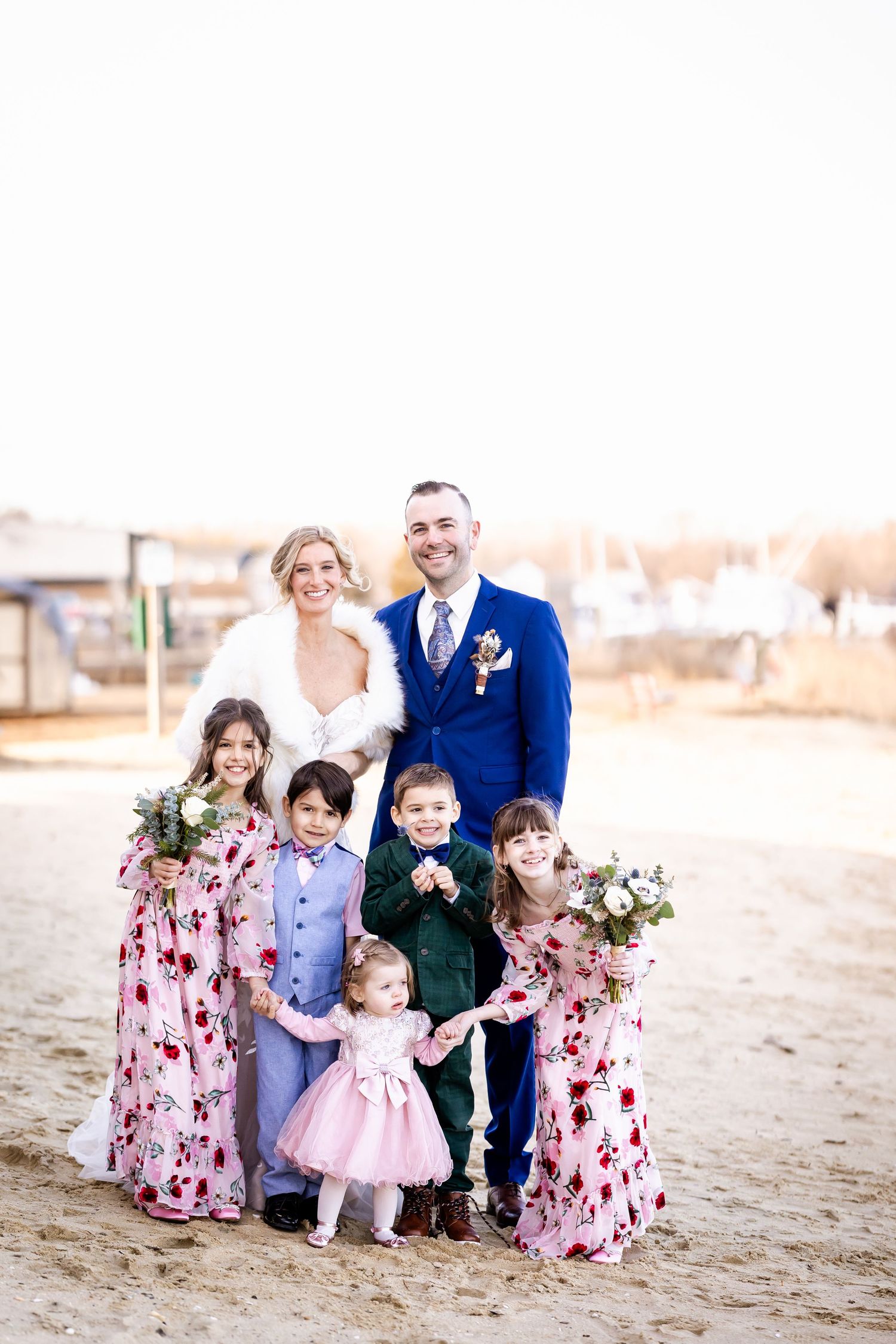 A family in coordinated outfits with floral dresses and formal wear poses together on a sandy beach at sunset.