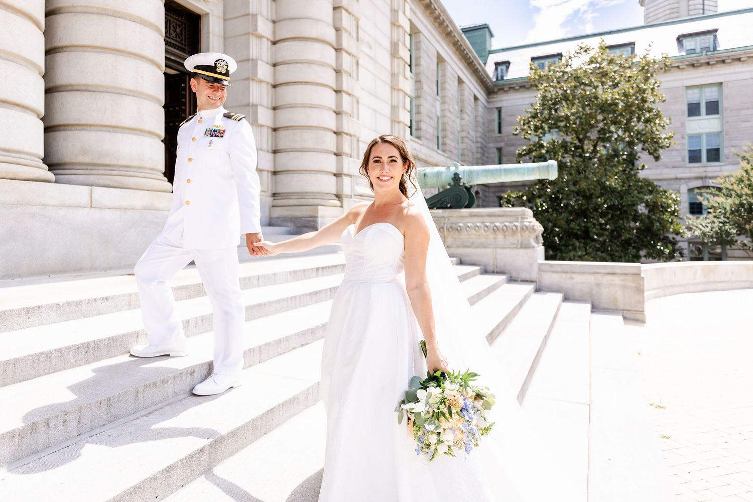 A bride in a white gown and naval officer in dress whites stand on grand marble steps of a historic building.