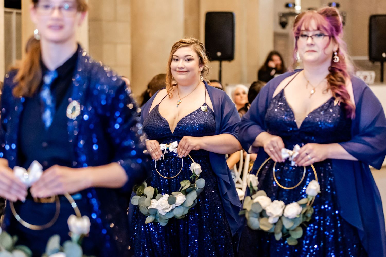 Three bridesmaids wearing matching navy blue sequined dresses with sheer overlays carry white bouquets.