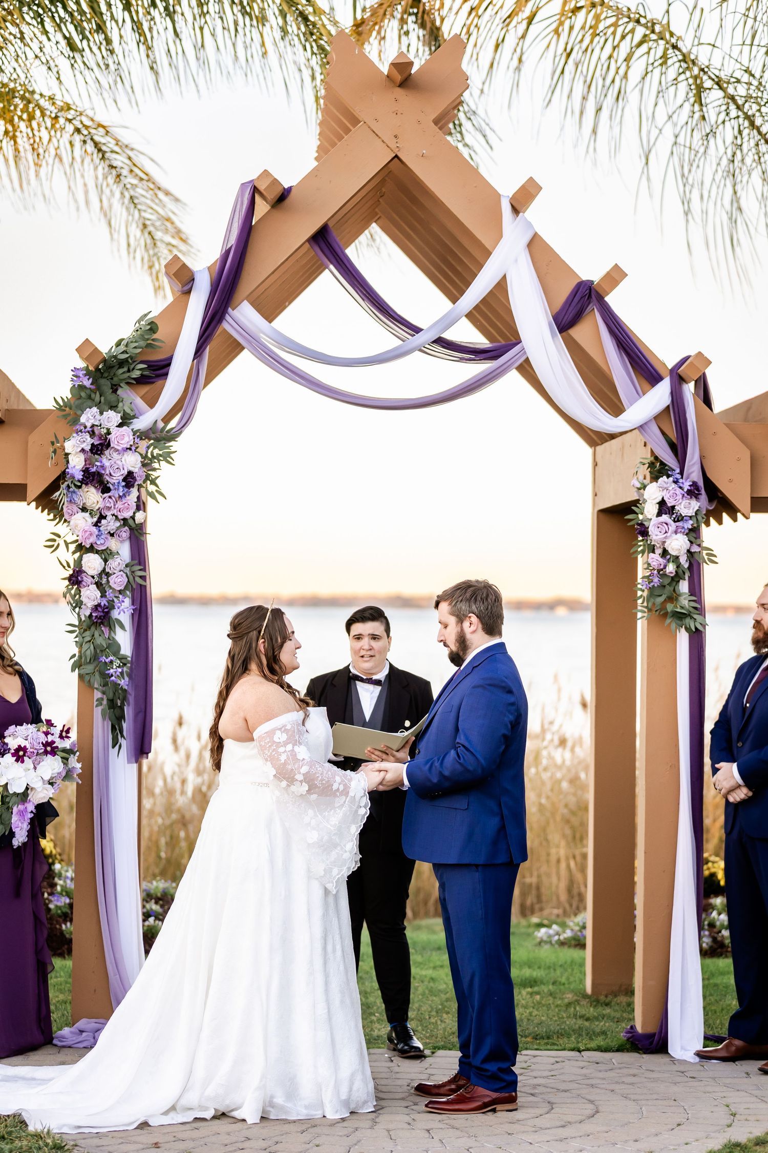 Wedding ceremony under wooden pergola with purple draping and florals at waterfront venue.